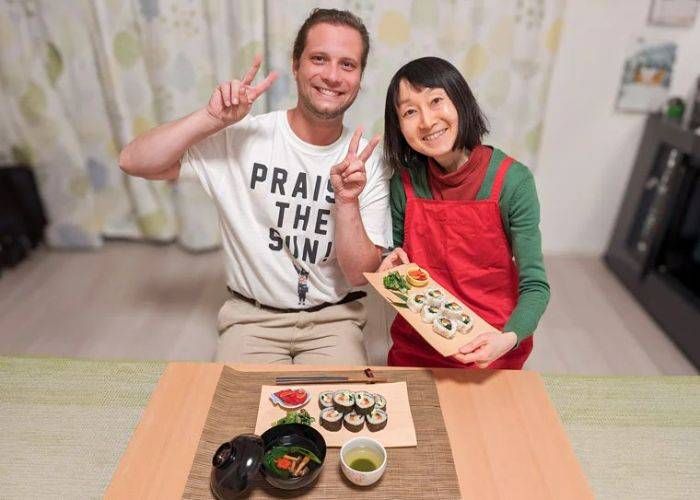 A host and a guest smile and show peace signs to the camera. In front of them, freshly made vegan sushi rolls.